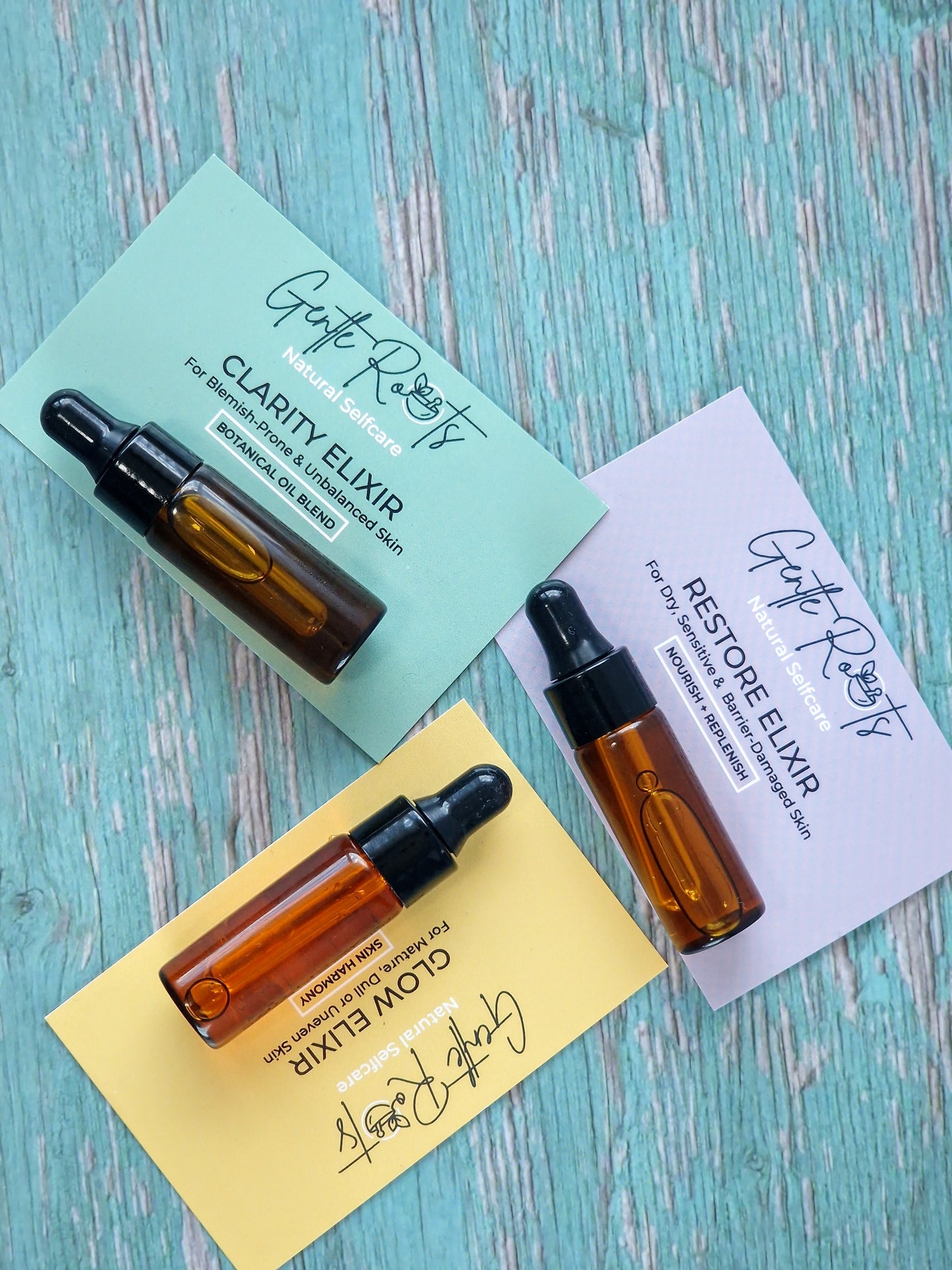 Three small amber bottles of the gentle roots mini elixirs collection with black caps on colorful cards against a textured blue background.