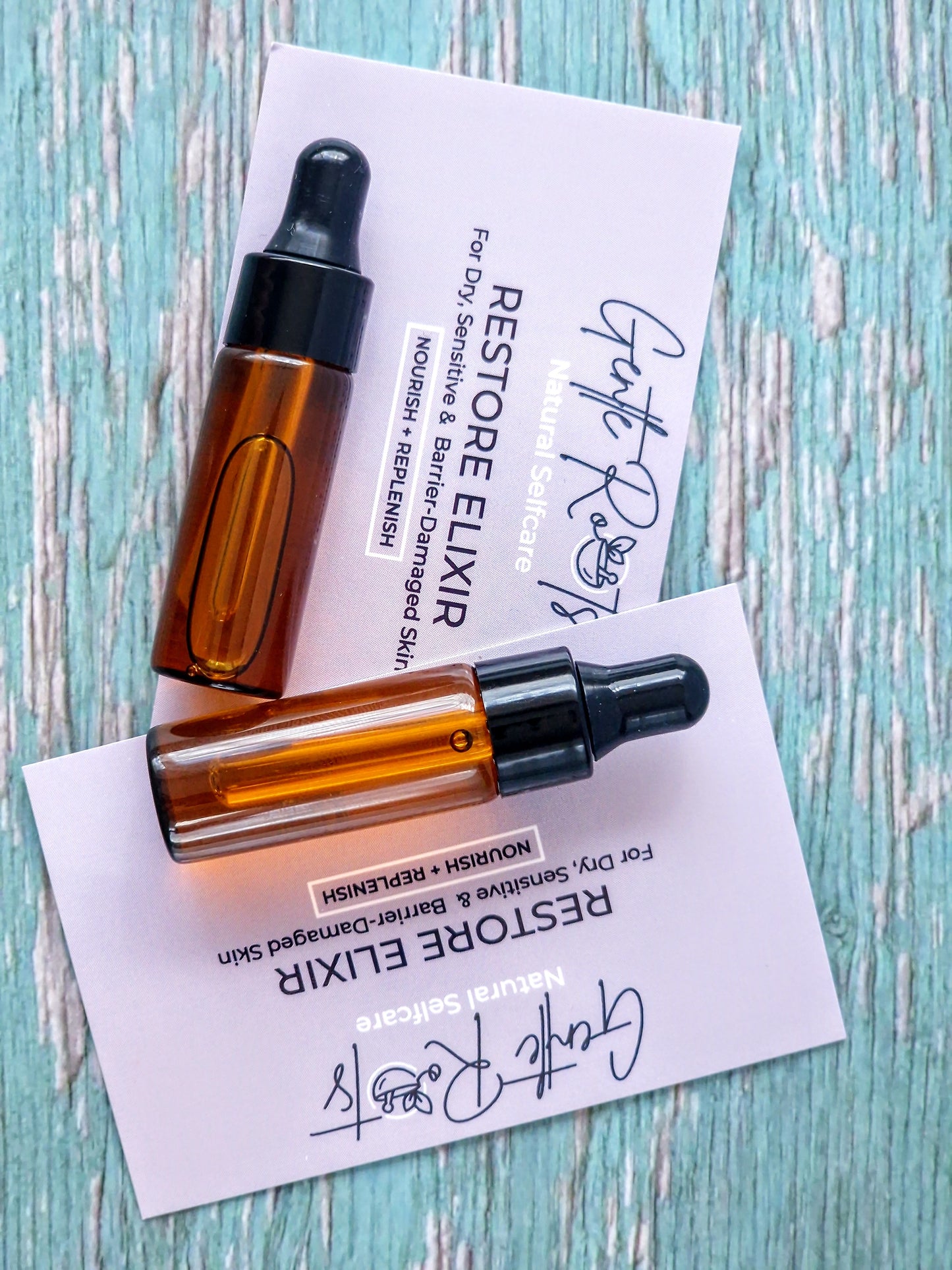 Two amber glass dropper bottles on a lilac card with 'Restore Elixir' text on a light blue wooden surface.
