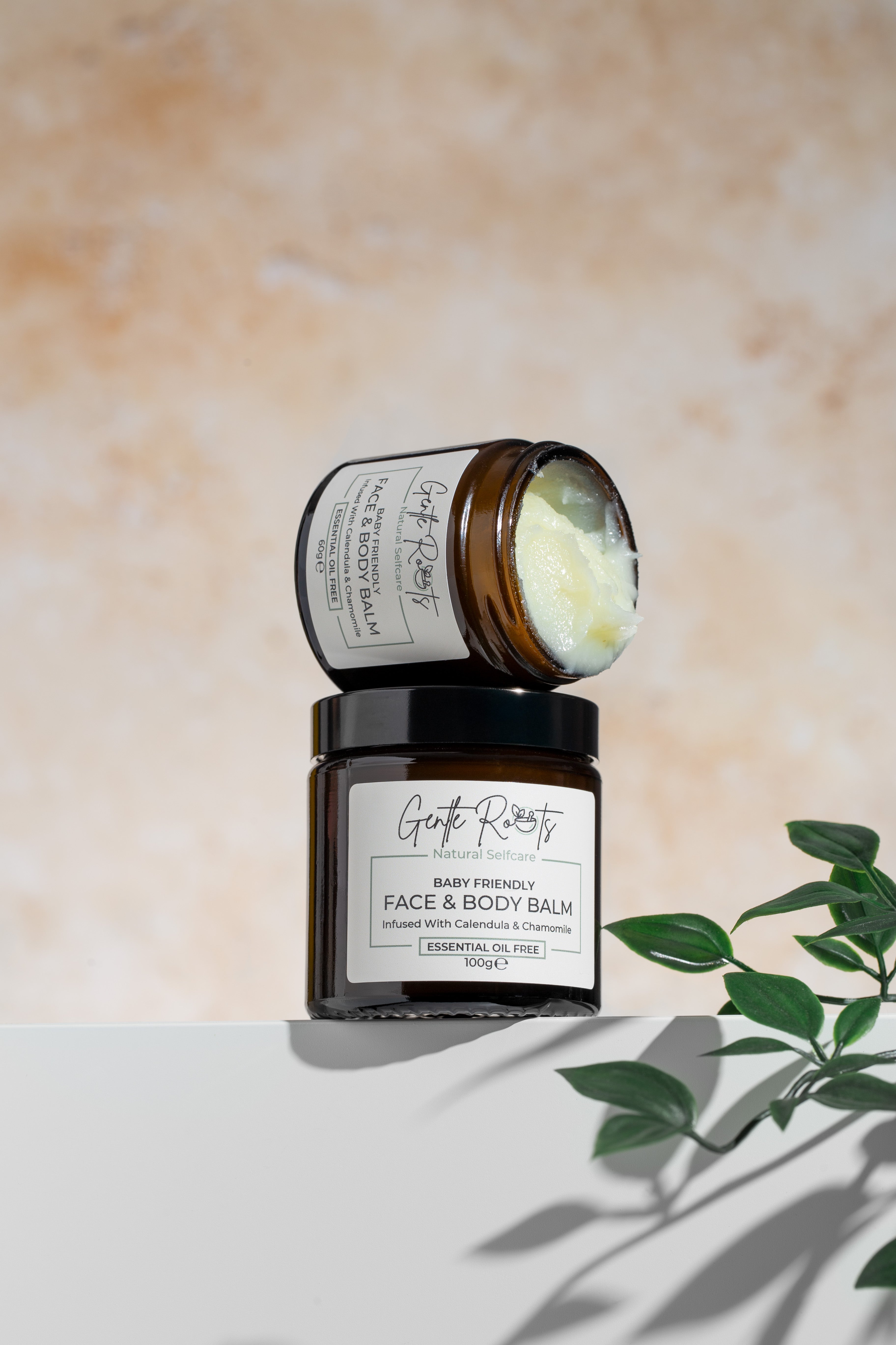 Gentle Roots Baby Friendly Tallow Balm in amber glass jars, open and stacked, with creamy balm visible, against a warm beige background and green foliage.
