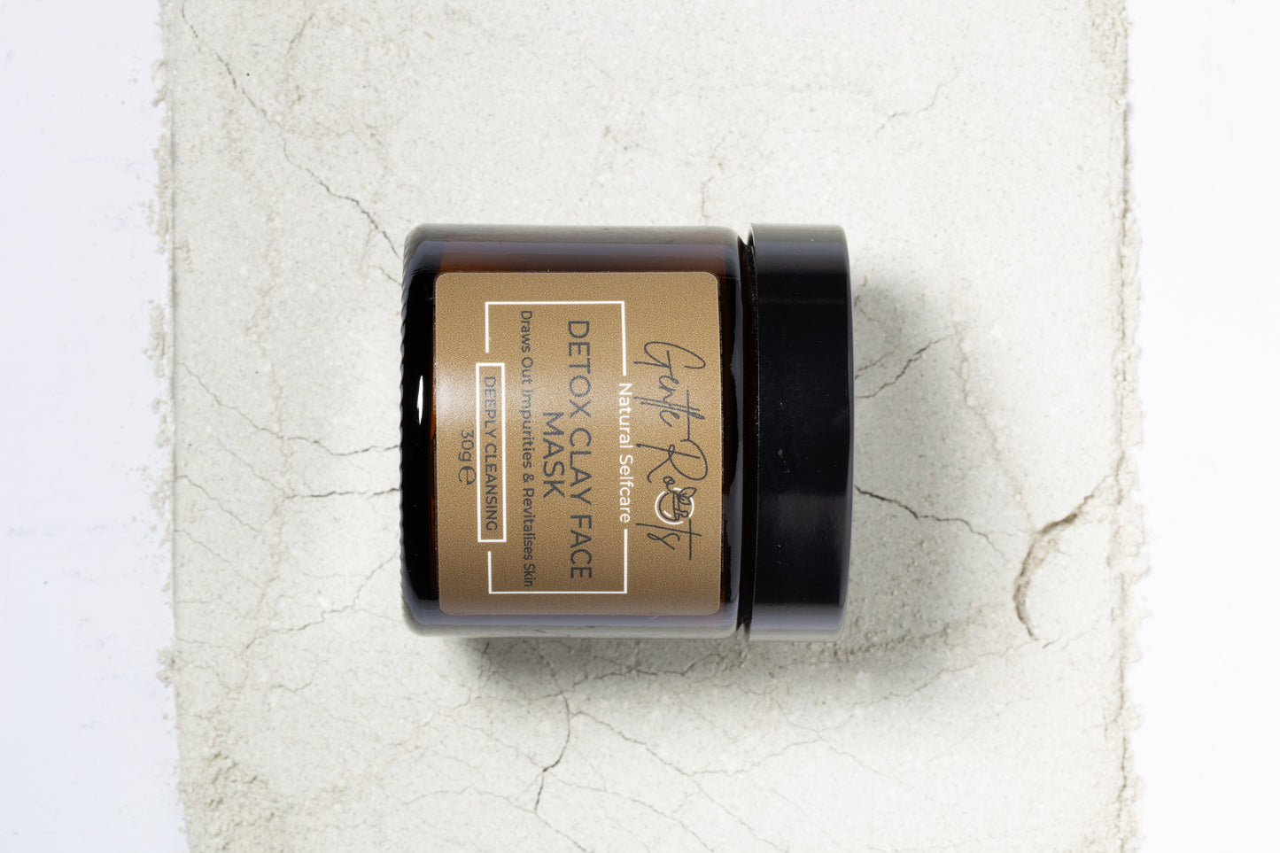 Gentle Roots Detox Clay Face Mask in amber glass jar on a background of powdered clay for a texture shot