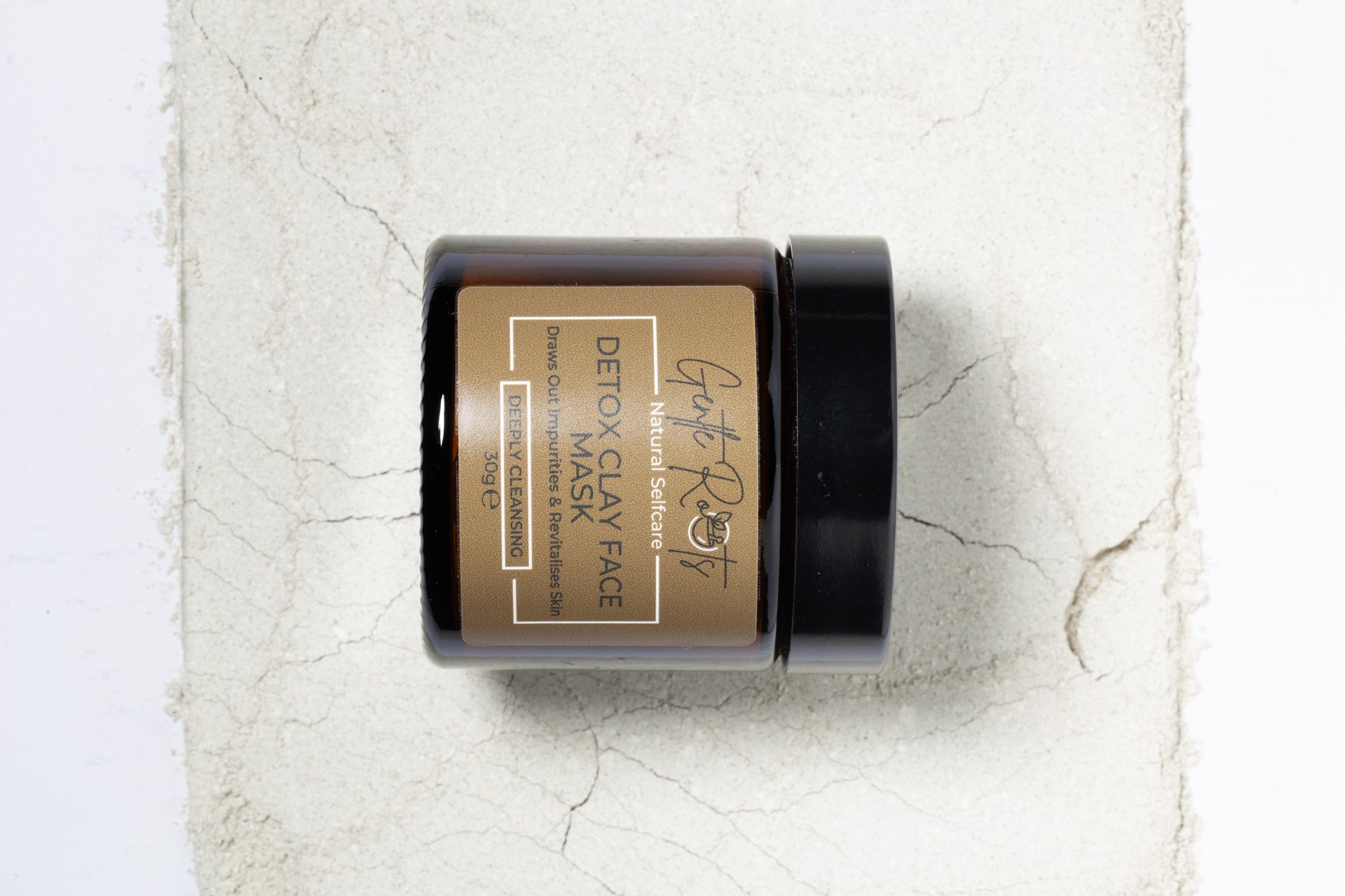 Gentle Roots Detox Clay Face Mask in amber glass jar on a background of powdered clay for a texture shot