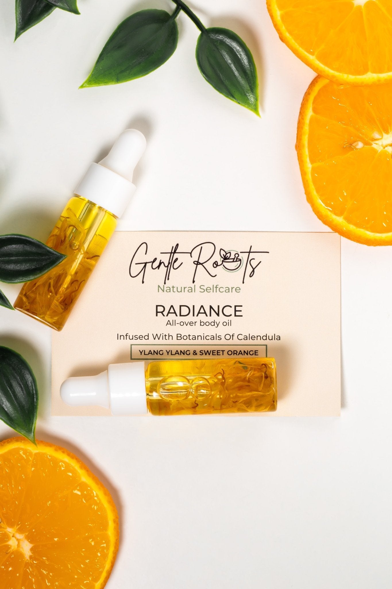 Gentle Roots Radiance Body Oil flat lay with fresh orange slices and ylang ylang – glow-boosting natural care.
